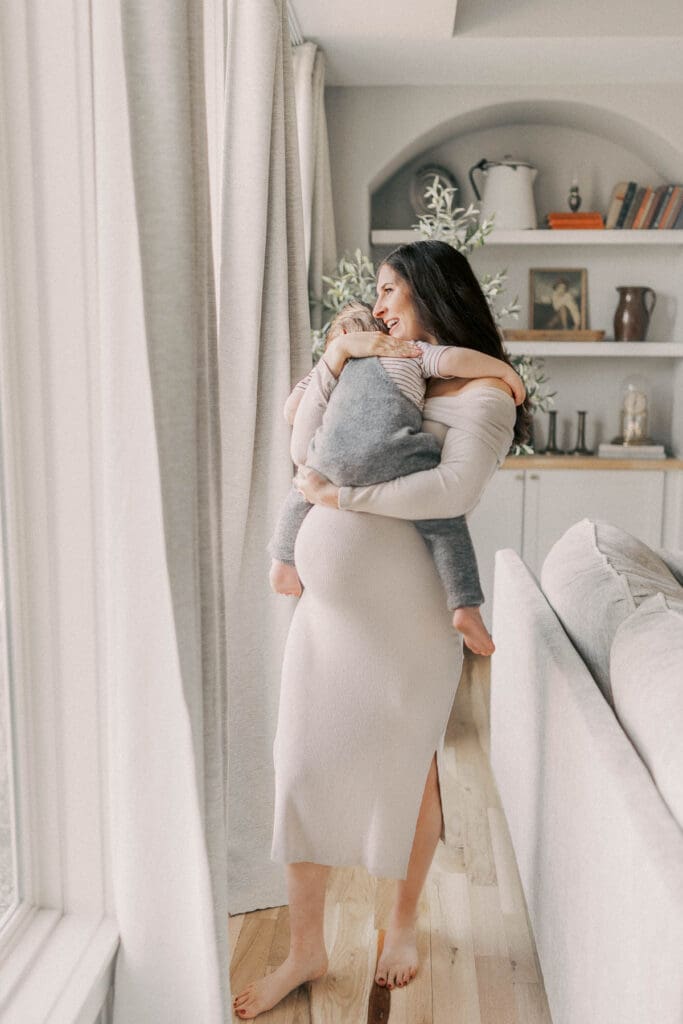 Mother and young son sharing a quiet embrace during in-home maternity photos in Kansas City, both wearing soft neutral tones in a light-filled bedroom.