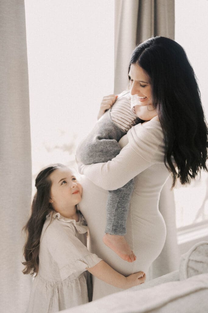 mother and two young children standing together in natural light during an in-home maternity session in Kansas City, styled in coordinated cream and beige clothing.