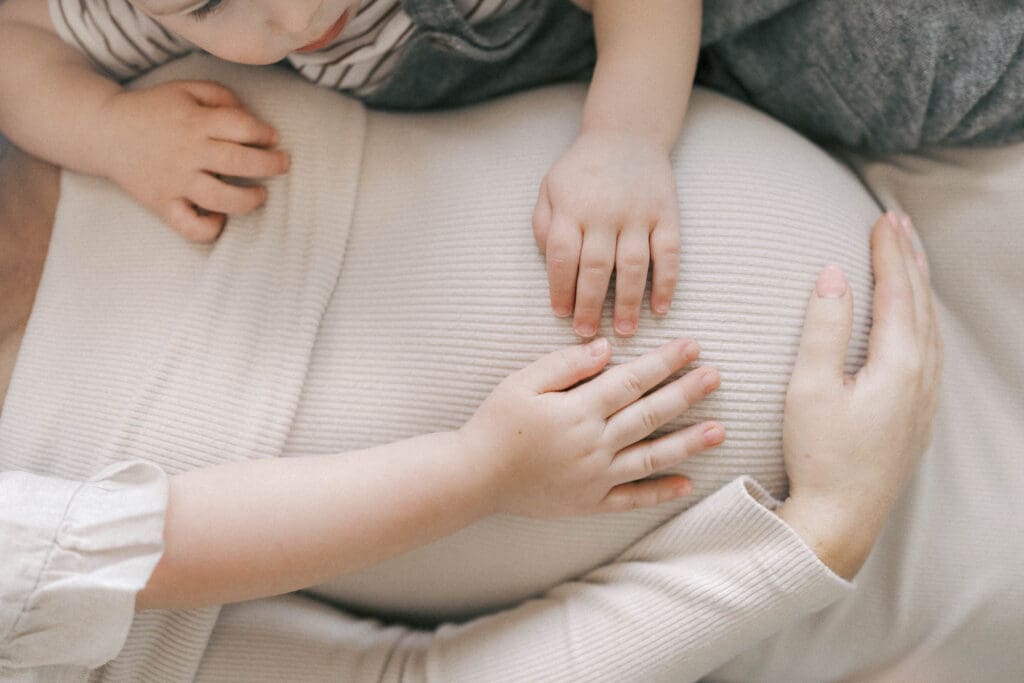 Pregnant mother wearing a fitted cream dress during an in-home maternity session in Kansas City, highlighting her baby bump in soft window light as young children touch her growing belly