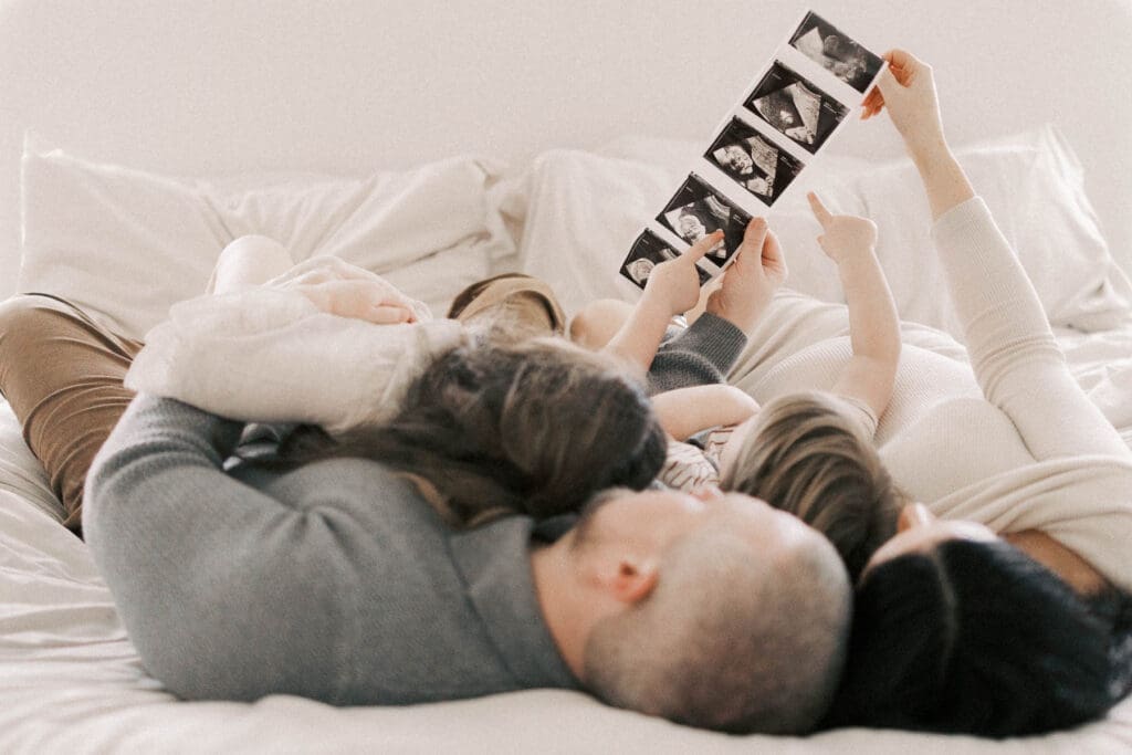 Mother, father, and their two young children sharing a quiet moment together as they look at ultrasound photos of their twin sisters during in-home maternity photos in Kansas City with a cozy, neutral aesthetic.
