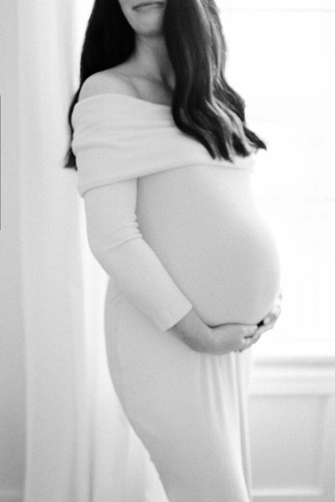 Expectant mother of twins stands near a window and looks over her shoulder during her in-home maternity photoshoot in kansas city