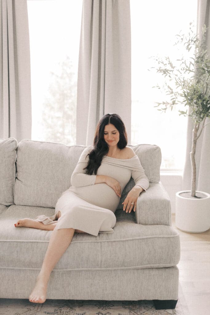 Expectant mother sitting on couch in a neutral-toned living room during in-home maternity photos in Kansas City, creating a calm waiting-for-baby feel.